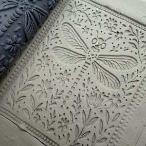 Peut inclure: Un rouleau à motifs gris foncé en argile placé à côté d'une plaque d'argile gris clair. Le rouleau et la surface de l'argile présentent un motif en relief représentant une grande libellule entourée de petites fleurs, feuilles et tiges.