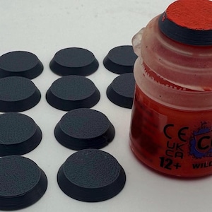 May include: Ten small, circular, black plastic bases with a textured top surface arranged in rows next to a translucent plastic paint pot containing bright red paint, featuring a red cap and text including "COLOR" and "WILD RIDER".