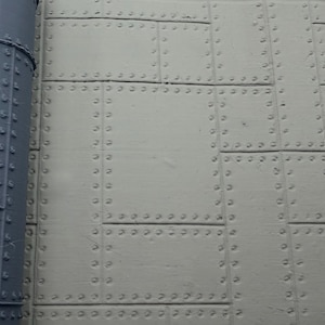 May include: A light grey surface composed of rectangular panels joined by rows of raised circular rivets, featuring a dark grey cylindrical pipe section on the left side that also displays a vertical row of rivets along its length.