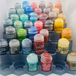 Könnte beinhalten: Ein abgestuftes graues Kunststoffregal mit zahlreichen kleinen Citadel Colour Farbtöpfen in verschiedenen Farben wie Rot, Blau, Grün, Gelb und Braun. Jeder Topf hat einen durchsichtigen Kunststoffdeckel und ein Etikett mit dem Markennamen.