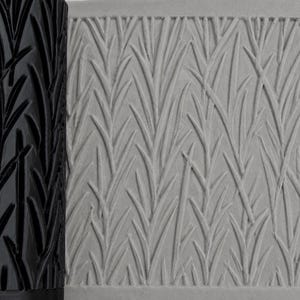 Könnte beinhalten: Eine schwarze zylindrische Teigrolle mit einem erhabenen, sich wiederholenden Gras- oder Schilfmuster, platziert neben einer hellgrauen Tonplatte, die dieselbe geprägte botanische Textur auf ihrer Oberfläche zeigt.