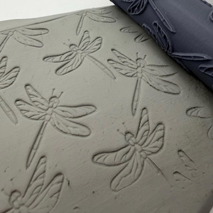 May include: A dark grey cylindrical rolling pin with a raised dragonfly pattern is pressed into a flat slab of light grey clay, leaving a repeating, indented impression of the insect across the surface of the malleable material.