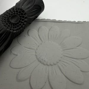 Könnte beinhalten: Eine schwarze, zylindrische Musterwalze mit Sonnenblumenmotiv neben einer flachen Platte aus grauer Tonerde, die mit demselben großen Sonnenblumenmuster geprägt wurde und die Textur sowie die Details der Blütenblätter zeigt.