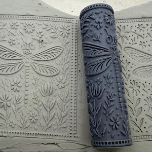 Könnte beinhalten: Zwei dunkelgraue zylindrische Tonwalzen mit geprägten Libellen- und Blumenmustern. Die Walzen liegen auf einer flachen hellgrauen Tonplatte und zeigen das eingeprägte Design einer Libelle, die von verschiedenen Blumen umgeben ist.