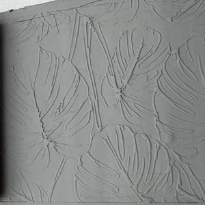 May include: A dark gray cylindrical clay rolling pin with a recessed monstera leaf pattern. The roller is positioned next to a flat, light gray slab of clay that displays a raised, embossed impression of the same monstera leaf design.