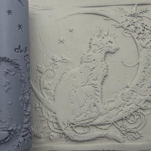 May include: A gray cylindrical clay rolling pin with a recessed pattern featuring a cat sitting on a crescent moon surrounded by floral vines and stars. The pattern is pressed into a flat slab of light gray clay, showing the embossed design.
