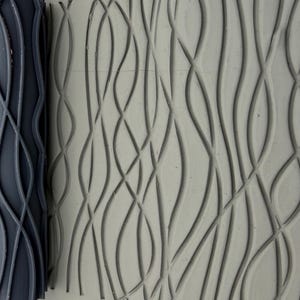 May include: A dark grey cylindrical rolling pin with a raised, repeating pattern of vertical, intersecting wavy lines. The tool is positioned next to a light grey, flat surface that displays the same embossed, flowing wave design in relief.