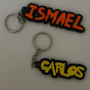 Puede incluir: Dos llaveros personalizados con base de goma negra y letras en relieve. Uno muestra el nombre ISMAEL en naranja brillante y el otro el nombre CARLOS en amarillo. Cada uno incluye una anilla y una cadena de metal de color plateado.