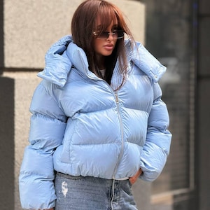 May include: A light blue puffer jacket with a high collar, a front zipper closure, and long sleeves. The jacket features a quilted design with horizontal baffles and is paired with blue denim jeans that have a distressed finish on the fabric.