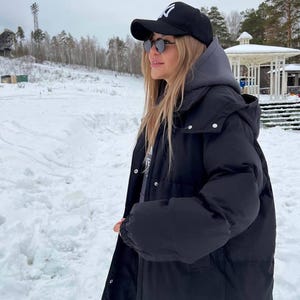 May include: A black baseball cap with a white logo, round dark-tinted sunglasses, a grey hooded sweatshirt, and a long black puffer coat with silver snap buttons, worn in a snowy outdoor environment with trees and a gazebo in the distance.