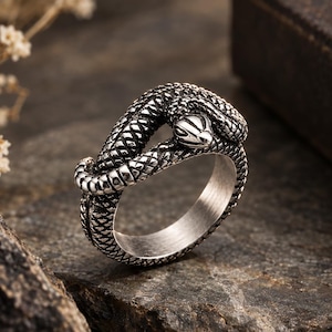 May include: A silver-toned metal ring shaped like a coiled snake. The band features a detailed, textured scale pattern covering the entire exterior surface, with the snake's head resting on the top of the band and its tail wrapping around the side.