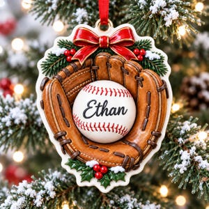 Peut inclure: Une décoration de sapin de Noël représentant un gant de baseball marron tenant une balle de baseball blanche avec des coutures rouges et le prénom Ethan inscrit en lettres noires. L'ornement est orné d'un nœud rouge, de sapin et de baies.