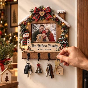 Può includere: Un portachiavi da parete in legno a forma di casa con cornice portafoto di una famiglia con cane, decorato con pupazzo di neve, albero di Natale, luci e un fiocco scozzese. Riporta la scritta The Wilson Family EST. 2020 e quattro ganci neri.