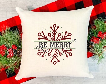 May include: A square, off-white decorative pillow with a snowflake design in burgundy and green, and the words "BE MERRY". The pillow is on a red and black plaid runner, with pine and berries on either side.