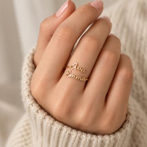 May include: A gold-toned ring featuring two stacked, open-ended bands, each displaying a name in a cursive script font. The top band reads Ava and the bottom band reads Emma. The metal has a polished finish and the design is delicate and thin.