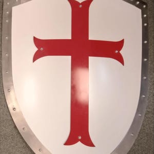 Puede incluir: Un escudo de tipo heráldico con una superficie blanca y un gran emblema de cruz roja centrado. El escudo presenta un borde metálico plateado asegurado con remaches visibles a lo largo de todo el contorno exterior, ofreciendo un aspecto reforzado.
