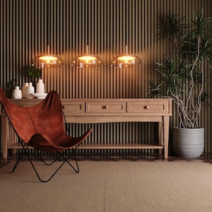 May include: A brown leather butterfly chair with a black metal frame positioned in front of a light wood console table. Three glass pendant lights hang above the table, which features small white vases and a potted plant to the right side.