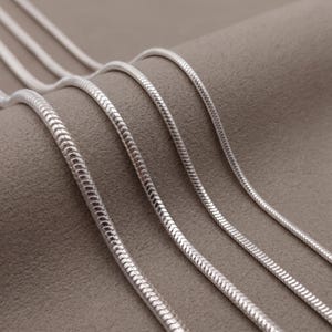 Könnte beinhalten: Vier silberfarbene Schlangenketten, die in parallelen Linien angeordnet sind. Jede Kette zeichnet sich durch ein glattes, flexibles und abgerundetes Design mit einem engen, ineinandergreifenden Gliedermuster aus, das eine durchgehende, glatte Oberfläche erzeugt.