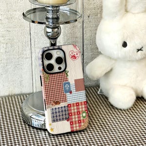 Puede incluir: Una funda de teléfono rectangular con un diseño de patchwork de cuadros vichy y escoceses en rojo, azul y blanco. La superficie incluye pequeñas ilustraciones de un corazón, una mariquita, una estrella y un pequeño animal.
