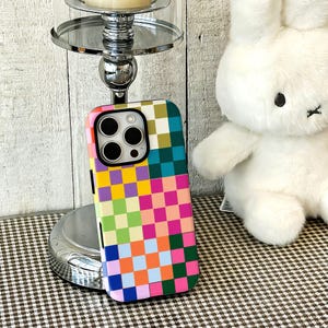 May include: A smartphone case featuring a vibrant, multi-colored checkerboard pattern with squares in shades of yellow, pink, orange, green, blue, and dark green. The case has a black camera cutout frame and covers the back of the mobile device.