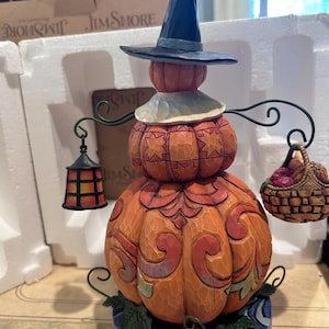 Peut inclure: Une figurine décorative en forme de citrouille portant un chapeau noir, avec une texture sculptée et des motifs orange, rouges et verts. Le personnage tient une lanterne et un panier de fruits, posé sur une base à motifs floraux.