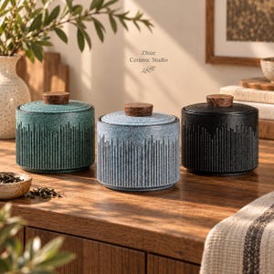 May include: Three cylindrical ceramic canisters with lids, featuring a textured vertical ribbed pattern. The canisters are colored in teal, light blue, and black, each topped with a small, round, brown wooden knob. Text reads Zhiee Ceramic Studio.