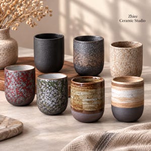 Puede incluir: Una colección de siete tazas de cerámica con texturas y acabados variados, incluyendo patrones en negro sólido, rojo moteado, verde, marrón y beige. El texto Zhiee Ceramic Studio es visible en la esquina superior derecha de la imagen.