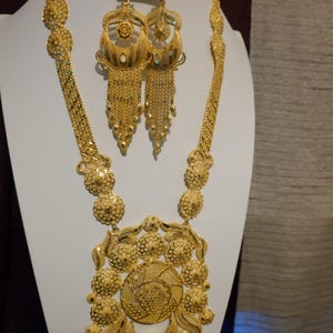 May include: A gold jewelry set consisting of a long necklace with a large circular pendant featuring floral patterns and dangling chains, a pair of matching drop earrings with intricate filigree work, and a matching gold ring with a crescent design.