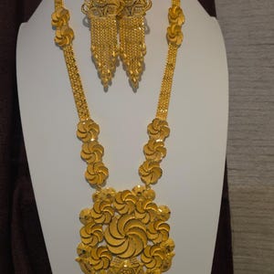 May include: A gold jewelry set consisting of a long necklace with floral links and a large circular pendant with dangling chains, matching drop earrings with intricate circular designs and fringe, and a matching gold ring on top.