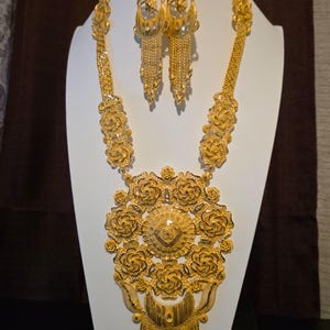 May include: A gold jewelry set consisting of a long necklace, a pair of drop earrings, and a ring. The necklace features a large, intricate floral pendant with a central textured disc and a dangling fringe. The earrings mirror the pendant design.