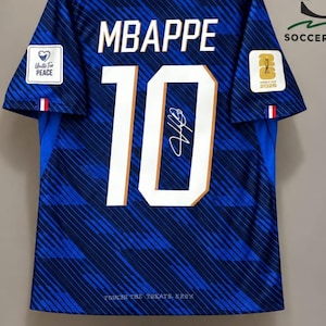 Puede incluir: Una camiseta de fútbol azul con cuello blanco, que muestra el nombre MBAPPE y el número 10 en blanco en la parte posterior. La camiseta tiene un patrón de rayas diagonales, una firma sobre el número y parches en las mangas. Un certificado enmarcado está debajo.