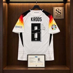 Könnte beinhalten: Ein weißes Fußballtrikot der deutschen Nationalmannschaft mit dem Namen KROOS und der Nummer 8 in Schwarz auf der Rückseite. Die Ärmel haben ein rot-gelb-oranges Farbverlaufsdesign mit weißen Streifen. Eine Unterschrift ist neben der 8 zu sehen.