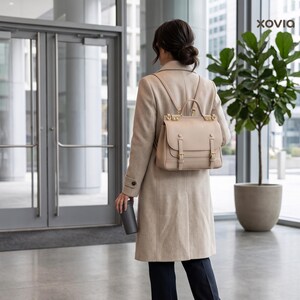 May include: A beige leather backpack with a top handle, two adjustable shoulder straps, and two front buckle closures. The bag features gold-tone hardware and a structured flap design. The brand name Xovia is visible in the top right corner.