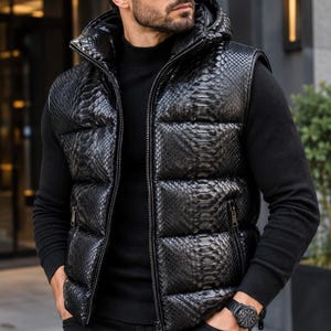 Peut inclure: Une doudoune sans manches noire avec un motif texturé en peau de serpent, une fermeture éclair intégrale sur le devant et une capuche. Le vêtement est porté sur un pull noir à manches longues, avec un pantalon noir et des lunettes de soleil.