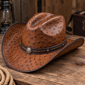 Puede incluir: Un sombrero de vaquero marrón con un patrón texturizado de piel de avestruz y una banda trenzada de color marrón oscuro. La banda está decorada con tachuelas de metal plateado y un adorno circular central con una estrella de cinco puntas.