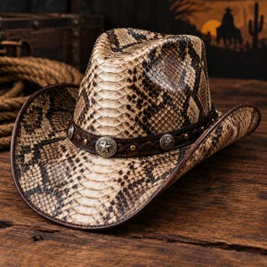 May include: A cowboy hat featuring a tan and dark brown snakeskin pattern across the crown and wide brim. A dark brown leather band encircles the base, decorated with braided leather details, small gold-tone studs, and silver-tone star conchos.
