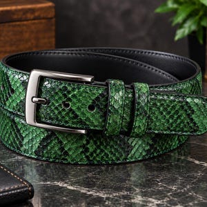 Puede incluir: Un cinturón con estampado de piel de serpiente en verde y negro y forro interior negro liso. El cinturón incluye una hebilla metálica rectangular en tono bronce y dos trabillas a juego de piel de serpiente verde para sujetar la punta.