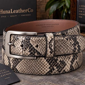 Puede incluir: Un cinturón de piel de pitón auténtica con un patrón natural de escamas en tonos beige, gris y negro. Cuenta con una hebilla de metal plateado cepillado y una trabilla de cuero a juego. El interior está marcado con Genuine Python Leather.