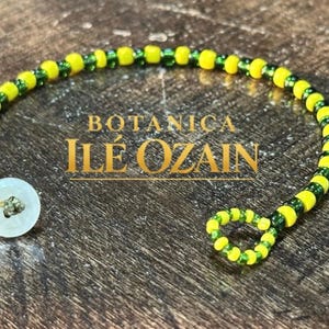 May include: A beaded bracelet featuring alternating yellow and green beads arranged in a circular pattern. The bracelet includes a white button closure and a small loop at the opposite end. The text BOTANICA ILÉ OZAIN is centered in the image.