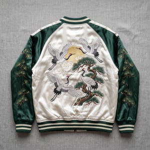 Puede incluir: Una chaqueta bomber de satén con un panel trasero color crema que presenta un diseño bordado de tres grullas, un sol dorado y un pino. Las mangas son de color verde oscuro con ramas de pino bordadas, y el cuello, los puños y el dobladillo son a rayas.
