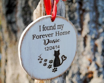 May include: A round, white wooden ornament with the text "I found my Forever Home Dasie 12/24/14". A black cat silhouette and paw prints are also featured. The ornament is hanging from a red ribbon.