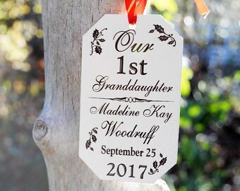 May include: A white tag with black text and floral accents, hanging from a tree. The tag reads "Our 1st Granddaughter Madeline Kay Woodruff September 25 2017". Red and orange ribbons are attached to the top of the tag.