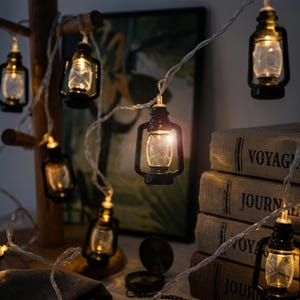 May include: A string of decorative fairy lights featuring miniature black lanterns with clear glass panes. Each lantern contains a warm glowing LED bulb. The lights are connected by a thin, flexible white wire, creating a soft illumination.