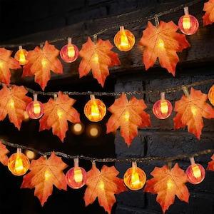Op de afbeelding: Een decoratieve lichtsnoer met afwisselend oranje en rode stoffen esdoornbladeren en kleine, ronde, gloeiende amberkleurige lampjes. De lampjes zijn gerangschikt in meerdere horizontale strengen, wat zorgt voor een warme verlichting.