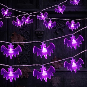 Pode incluir: Uma corrente de luzes LED roxas em forma de morcegos. Cada morcego roxo translúcido possui uma fonte de luz central que brilha intensamente. As luzes estão conectadas por um fio branco fino e flexível, criando um conjunto decorativo.