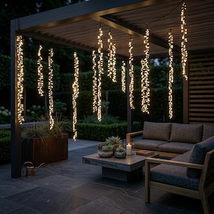Op de afbeelding: Een reeks verticale lichtsnoeren met warmwitte ledlampen die aan een donkere metalen pergolastructuur hangen. De lampjes zijn in meerdere strengen gerangschikt en creëren een gloeiend effect boven een terras met buitenmeubilair en planten.