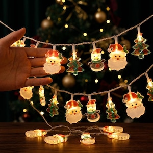 May include: A string of LED Christmas lights featuring translucent, illuminated figures of Santa Claus, snowmen, and pine trees. The decorative light strand is connected by a thin, clear wire, with several figures glowing warmly against a dark background.