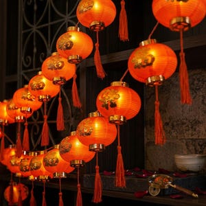 Op de afbeelding: Een lichtsnoer met gloeiende oranje papieren lantaarns voorzien van goudkleurige decoratieve patronen. Elke bolvormige lantaarn hangt aan een koord en heeft een bijpassend oranje kwastje aan de onderkant voor een warme verlichting.