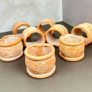 May include: A set of eight terracotta napkin rings, each featuring a cylindrical shape with a raised geometric pattern around the exterior. The rings have a warm, light orange hue and a smooth, matte finish, showcasing a consistent design.