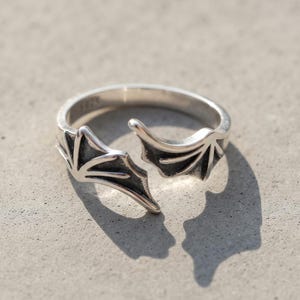 Puede incluir: Un anillo abierto de tono plateado que presenta dos motivos simétricos de alas de murciélago que se encuentran en el centro. El metal tiene un acabado pulido en los bordes elevados y una textura oscura y hundida en los detalles de las alas.